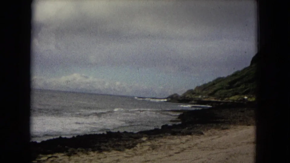 1960s 8mm coastal landscape with overcast sky, low waves, dark rocks, sand, and a green hillside at right.