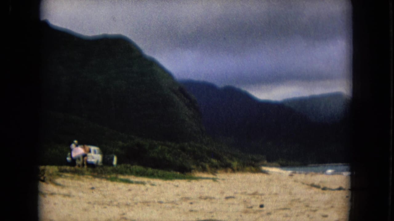 1967 New Mexico home-movie frame with a light-colored car and people near the beach, backed by mountains and water.