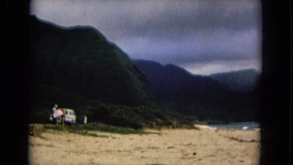 1967 New Mexico home-movie frame with a light-colored car and people near the beach, backed by mountains and water.