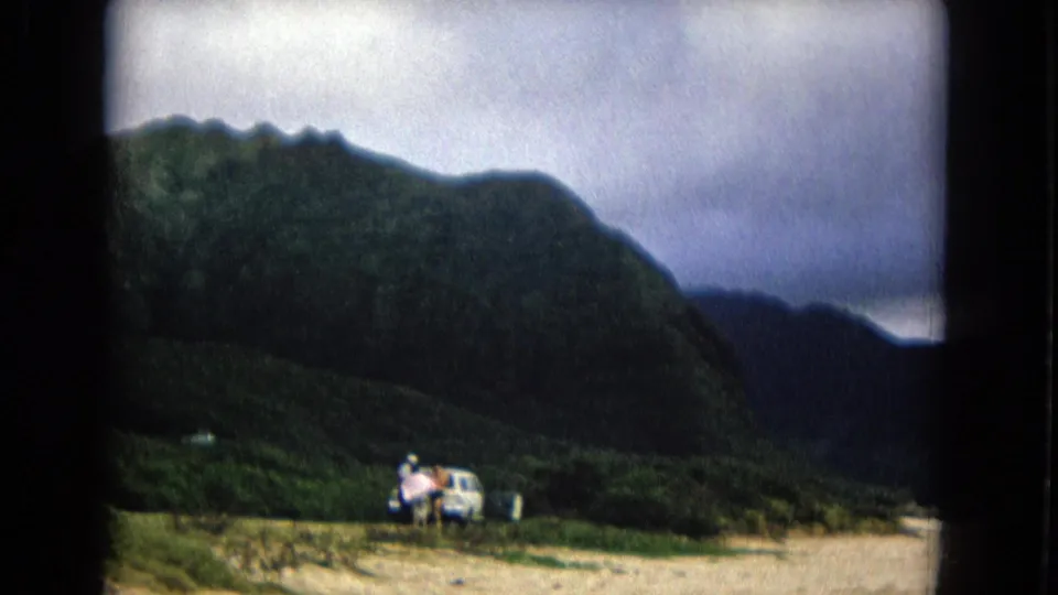 1967 New Mexico 8mm frame showing people near a parked car on sandy ground below dark mountain slopes.
