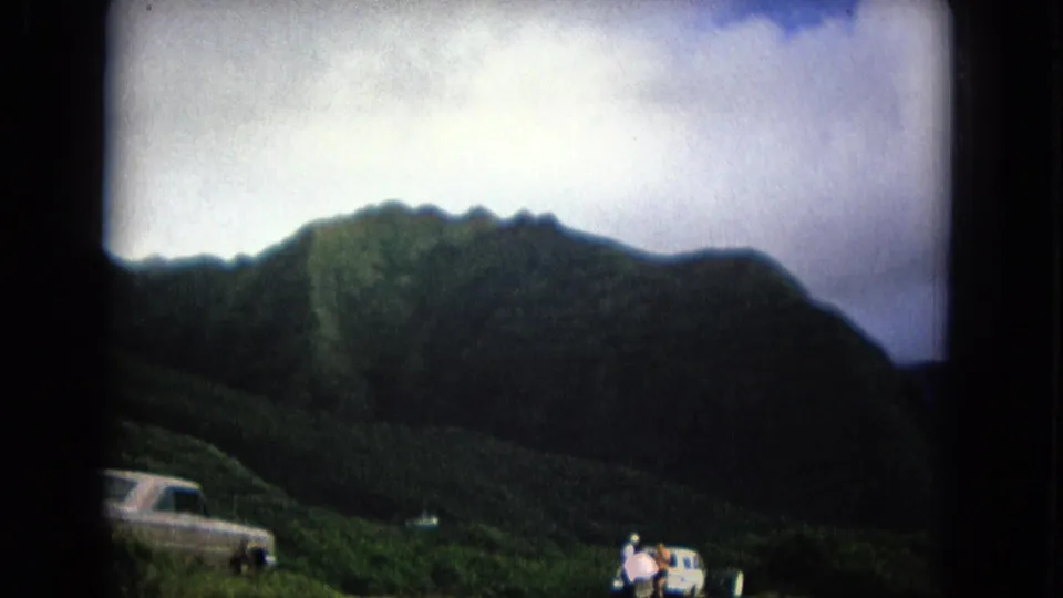 1967 New Mexico frame with a mountain ridge, cloudy sky, and a partially visible car and people along the lower edge.