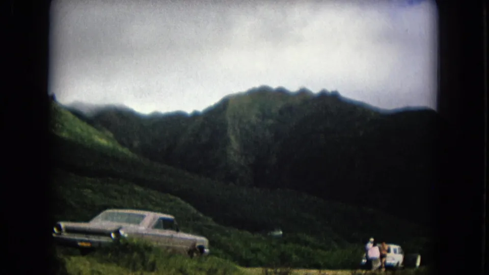 1967 New Mexico 8mm frame showing two classic cars stopped below green mountains with people gathered near the smaller vehicle.