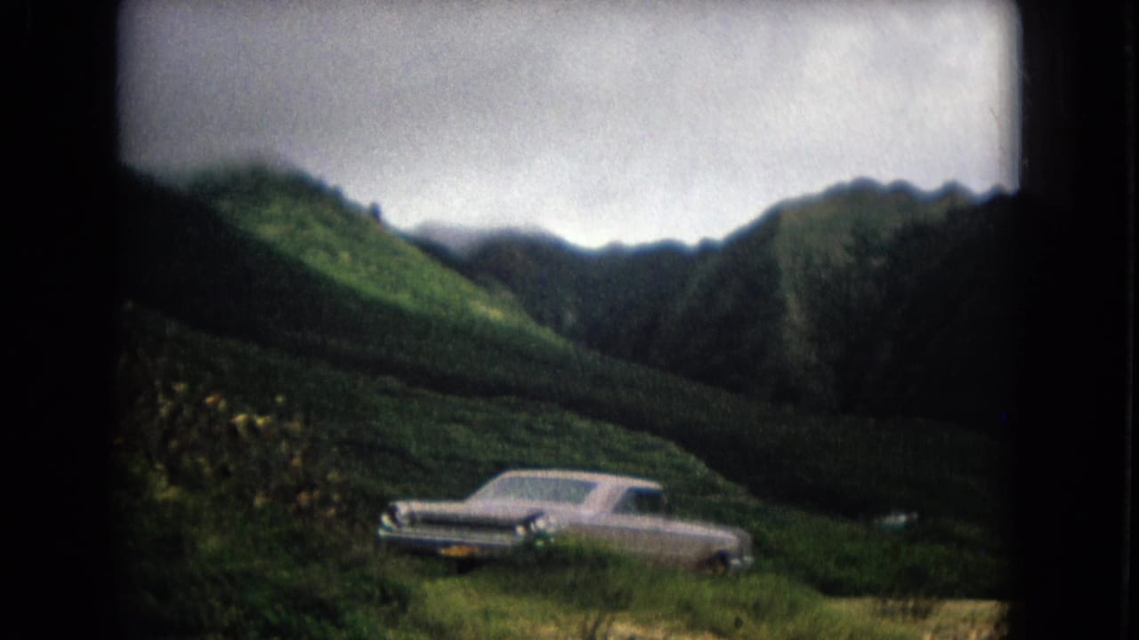 1967 New Mexico home-movie frame with a light-colored sedan parked near flowering vegetation under cloudy mountain ridges.