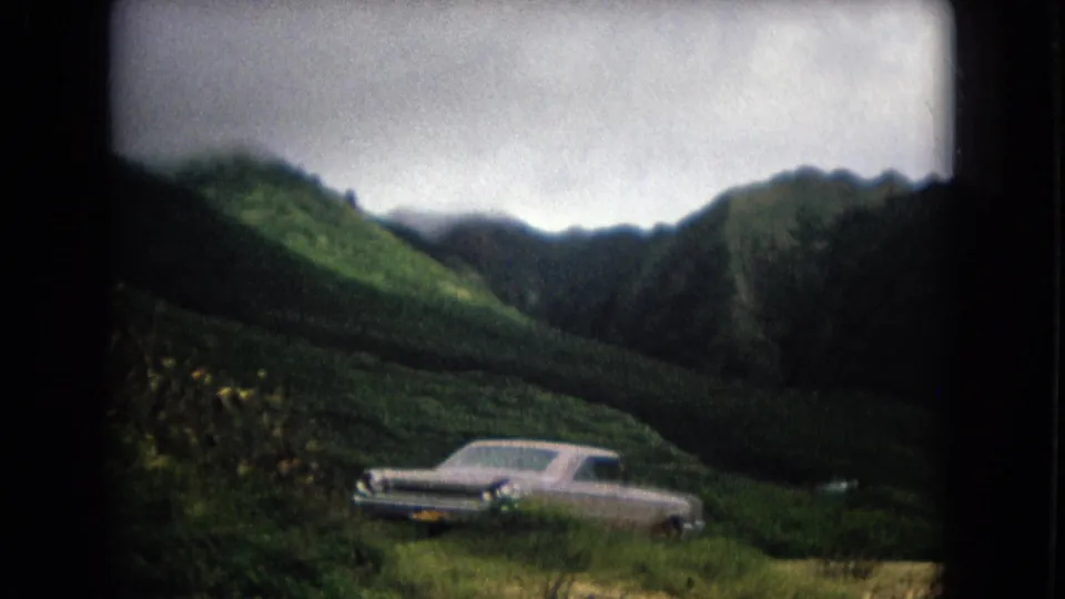 1967 New Mexico home-movie frame with a light-colored sedan parked near flowering vegetation under cloudy mountain ridges.