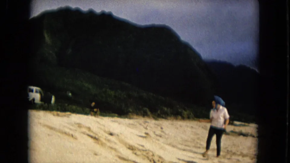 1967 New Mexico 8mm frame showing a person walking on sandy ground below dark green mountains, with a small vehicle in the background.