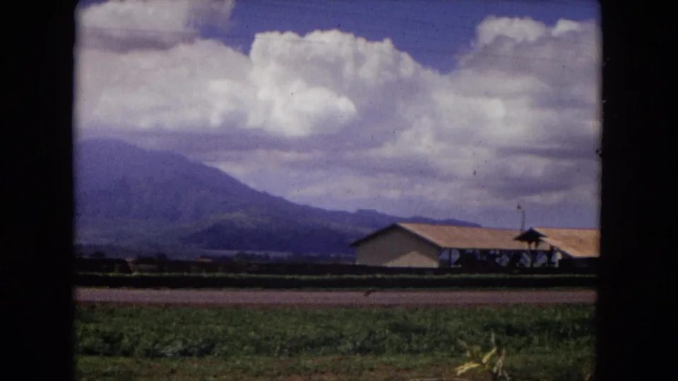 1967 New Mexico 8mm frame showing green fields, a low roofed building, distant mountains, and large white clouds from a moving vehicle.