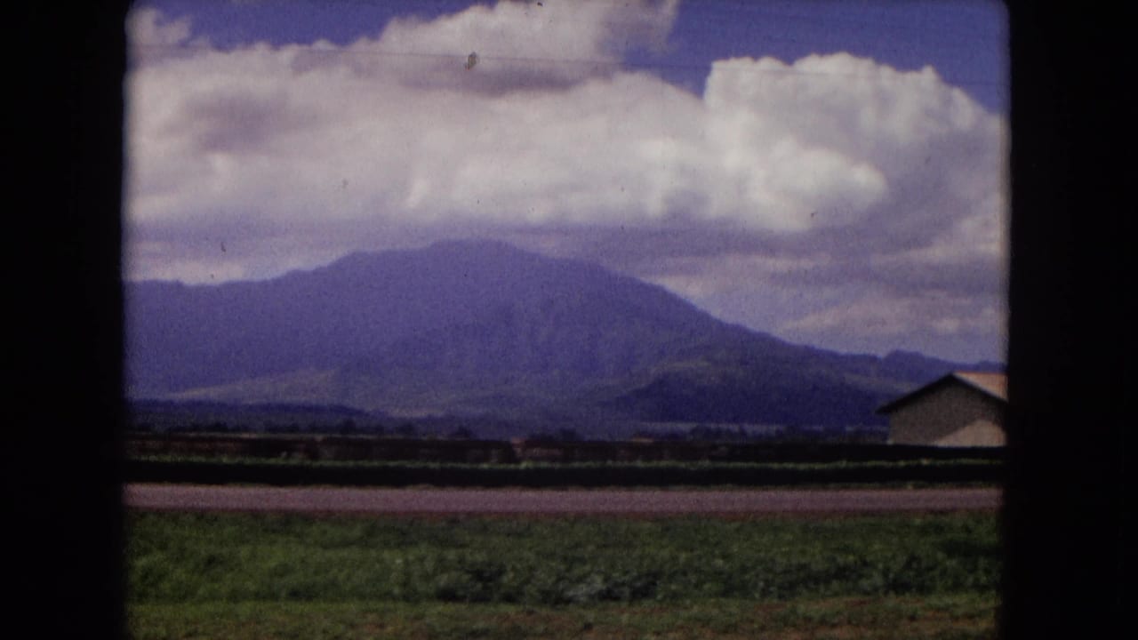 1967 New Mexico landscape frame with fields, a mountain range, large clouds, blue sky, and a partial low building at the right edge.