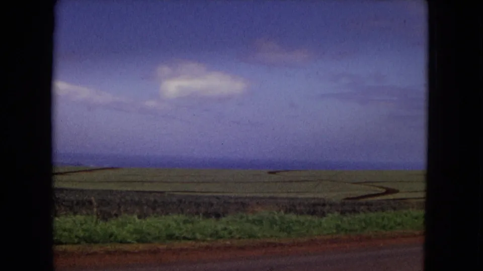 1967 New Mexico landscape frame showing flat fields, a reddish roadside edge, blue sky, and a small cloud near the horizon.