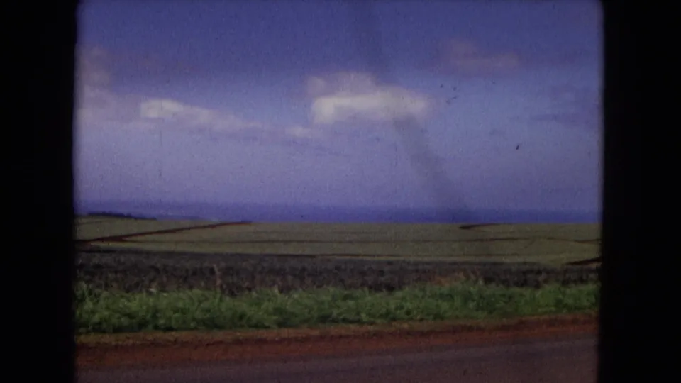 1967 New Mexico 8mm road-view frame showing green fields, reddish roadside soil, distant mountains, and a broad cloudy sky.