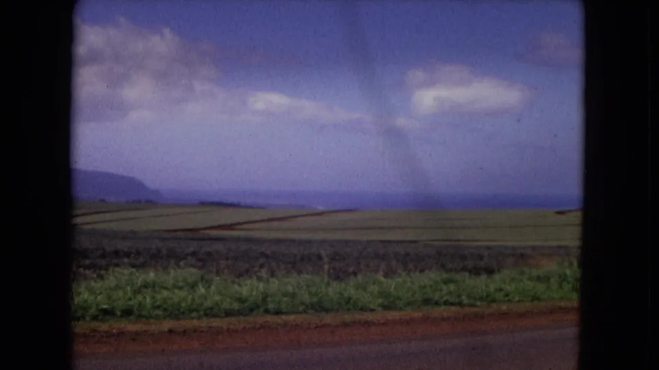 1967 New Mexico countryside frame with neat crop fields, a low mountain ridge, blue sky, clouds, and 8mm frame edges.