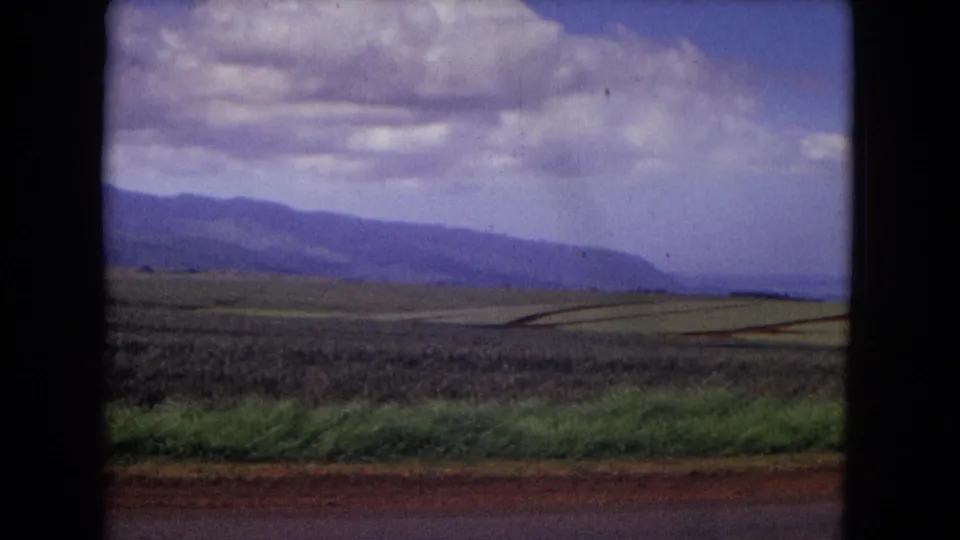 1967 New Mexico roadside landscape with patchwork fields, a dark foreground band, distant mountains, and textured clouds.