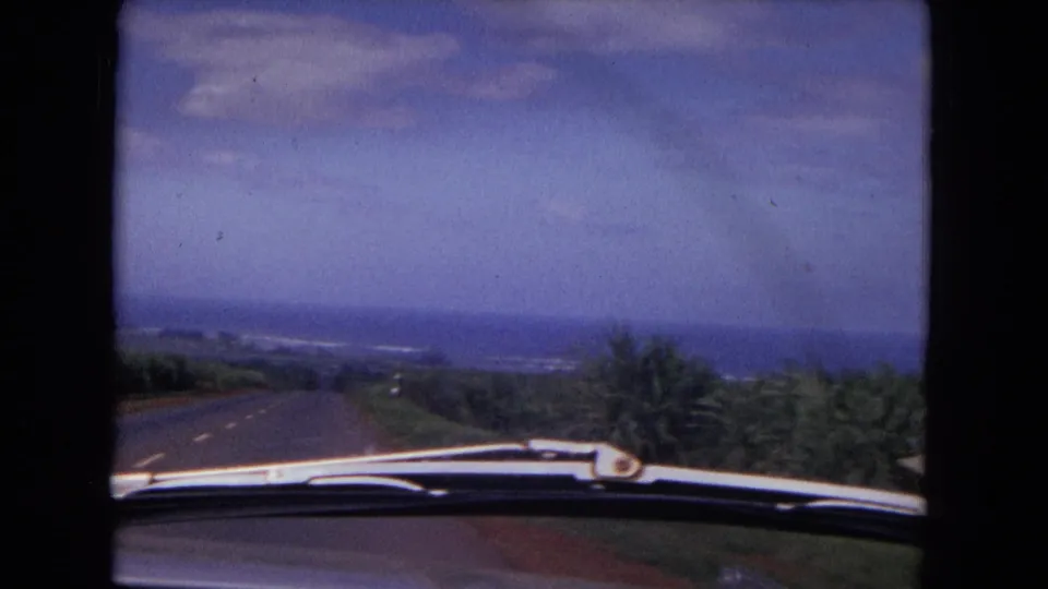 1967 8mm frame looking over a car hood down a paved road bordered by greenery, with a distant horizon under blue sky.