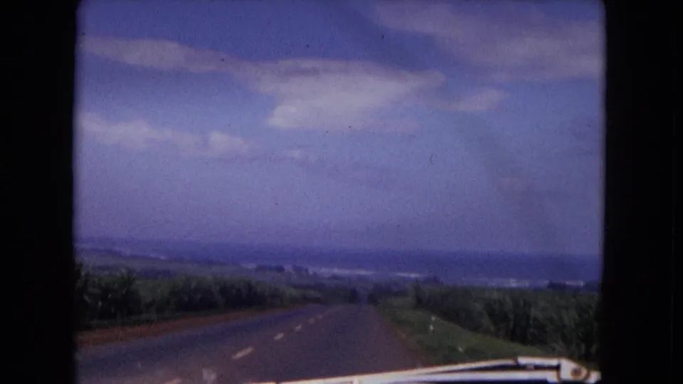 1967 8mm travel view over the car hood as the road descends through low vegetation toward distant blue water or haze.