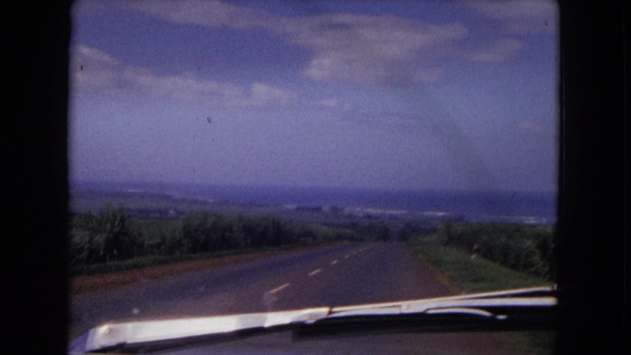 Road seen over a car hood with vegetation, distant hills, and a cloudy blue sky.