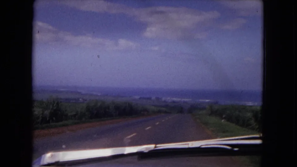1967 8mm travel view from inside a moving car, looking down a paved road toward distant hills and a hazy horizon.