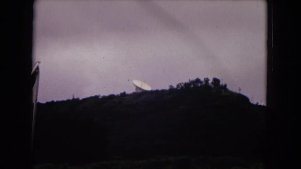 Hilltop view on vintage 8mm film showing a dish-like structure silhouetted near trees under a pale sky.