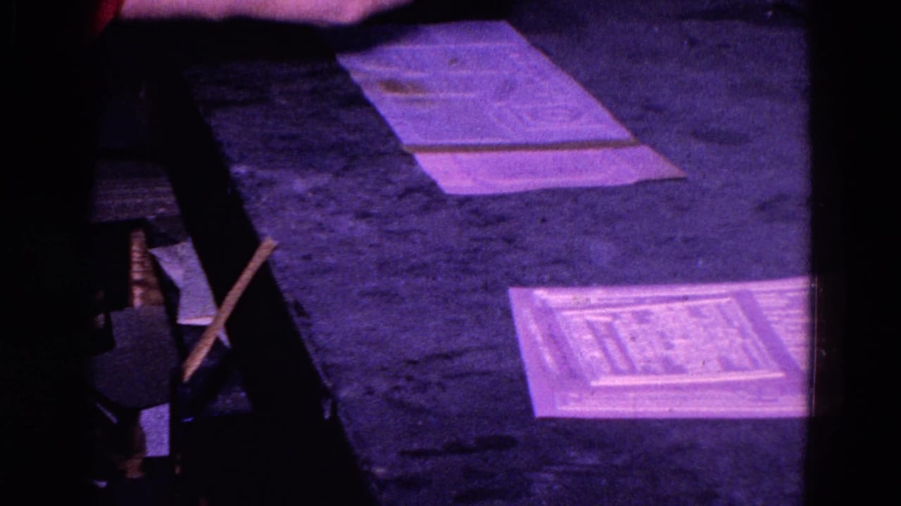 1967 Trenton home-movie frame showing papers on a dark desk or tabletop under purple low light.