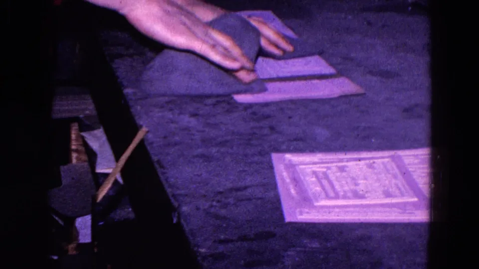 Close 8mm frame of a hand rubbing a cloth over a rectangular paper on a dark surface, supported as 1967 Trenton footage.