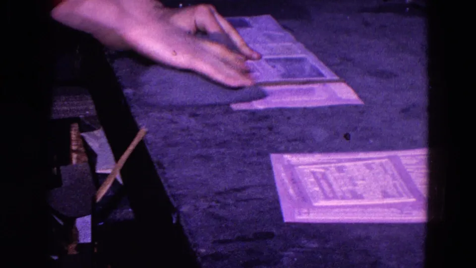 Hands and cloth pass over stacked or overlapping papers on a dark tabletop in purple-tinted 8mm home-movie footage.