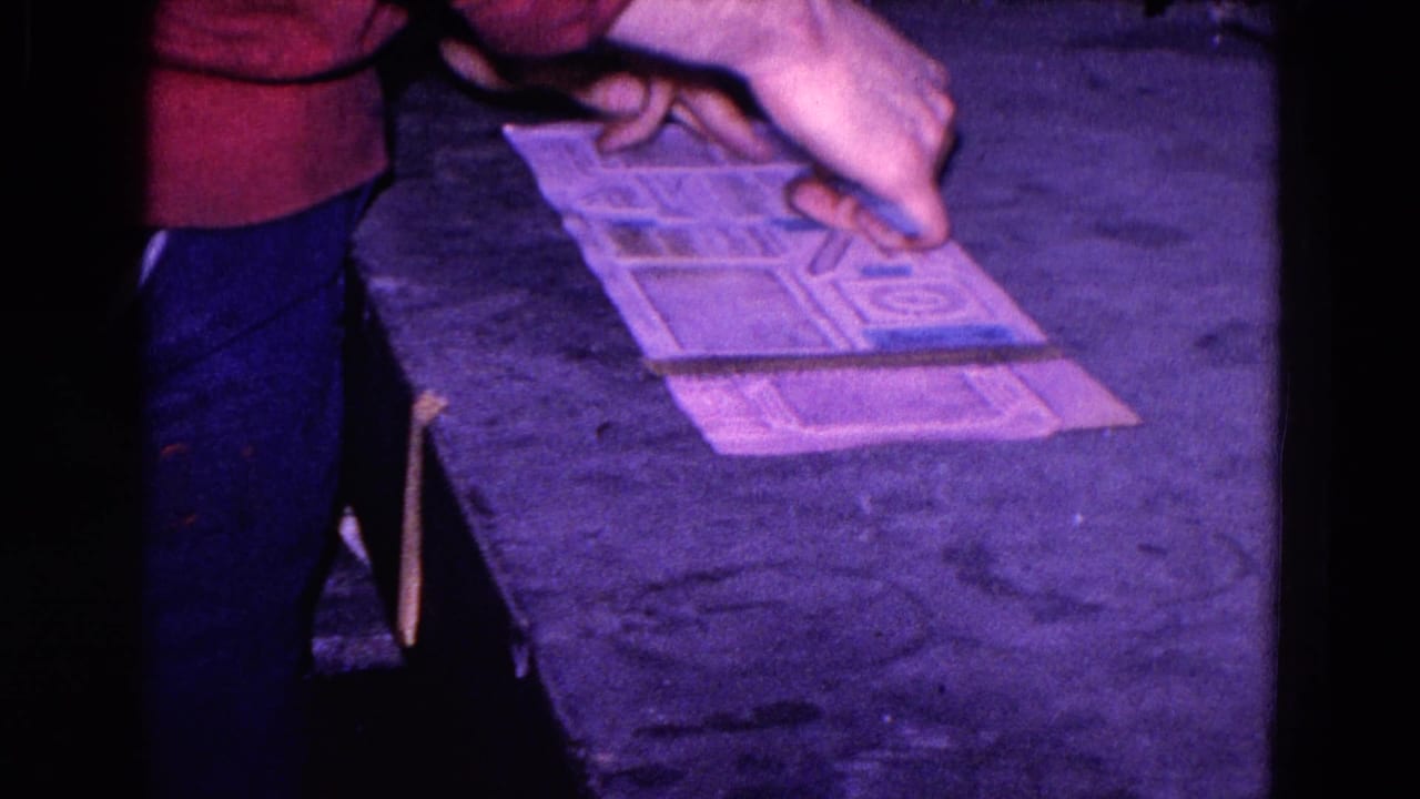 A person in a red sweater steadies a geometric paper template on a dark tabletop in the Trenton 1967 home-movie clip.