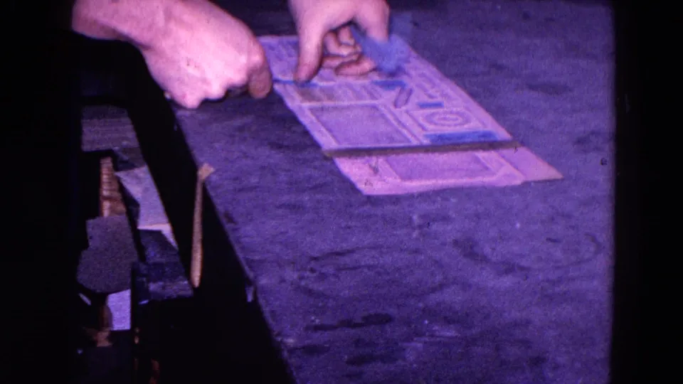 In 1967 Trenton factory footage, a worker's hands apply blue stickers to a printed sheet on a dim workbench.