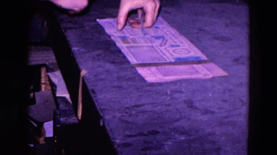 A close view shows hands arranging blue stickers on a printed paper sheet in the Trenton factory setting.
