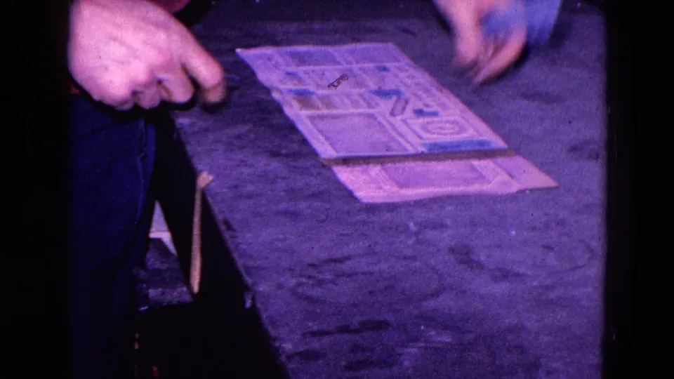 The worker's hand pauses beside the printed sheet as blue stickers remain arranged on the paper in the 1967 factory scene.