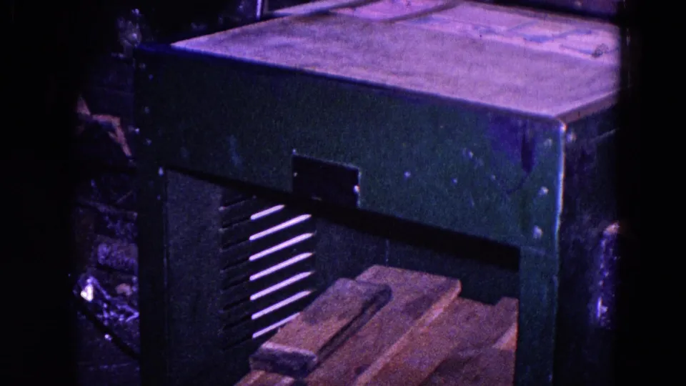 A green metal cabinet or machine sits near the workbench, with wooden blocks visible below in the Trenton industrial setting.