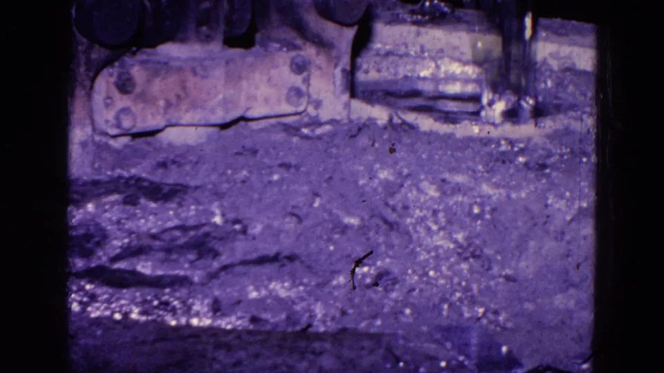1967 Trenton 8mm close-up of a rough foundry surface beneath dark machine parts and a small bright metal area, with visible dust and aged color shift.