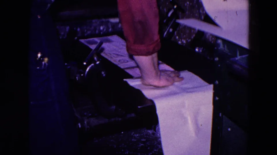 1967 Trenton industrial frame showing a worker's bare foot on white paper beside dark machinery, red pants, and a tray-like surface.