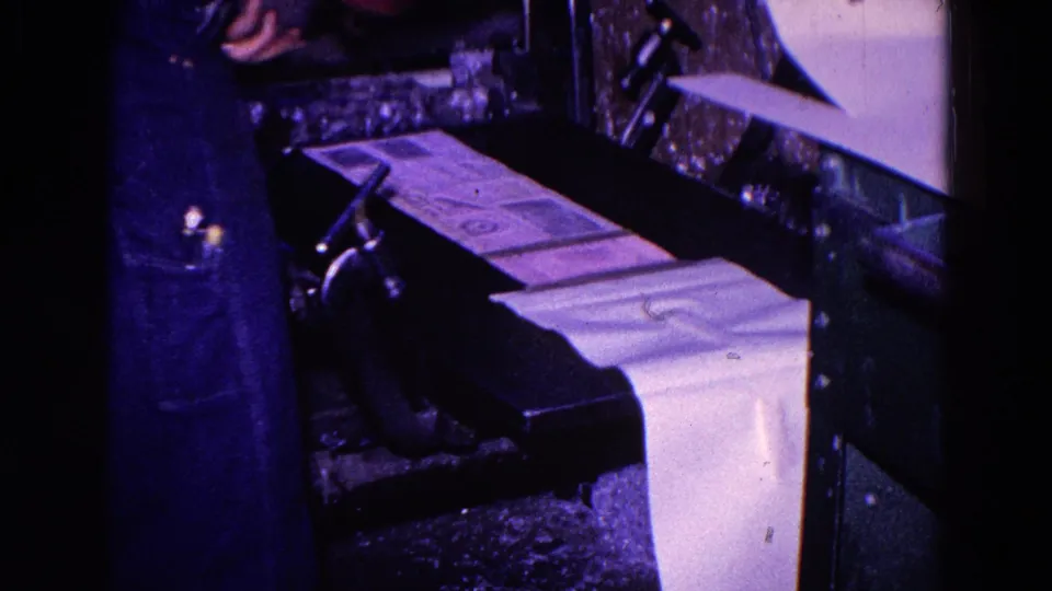 In 1967 Trenton, New Jersey footage, a worker in denim stands near a press with printed sheets feeding across the machine.
