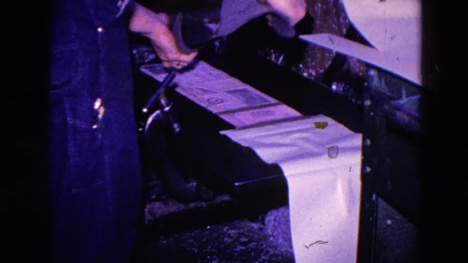 A worker reaches over the press as printed sheets continue through the machine in the Trenton printing plant footage.