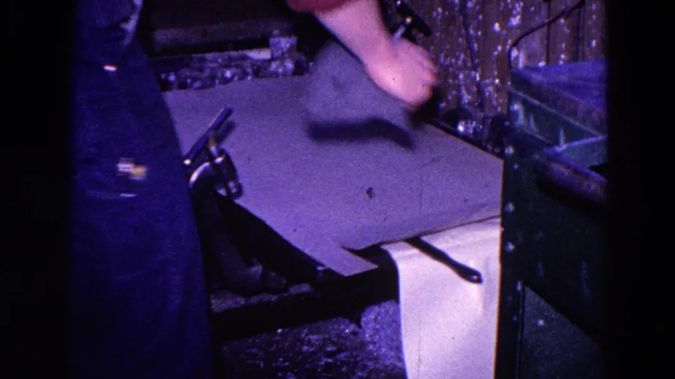 The worker lifts a broad sheet of paper beside the press, partially obscuring the printed feed below.
