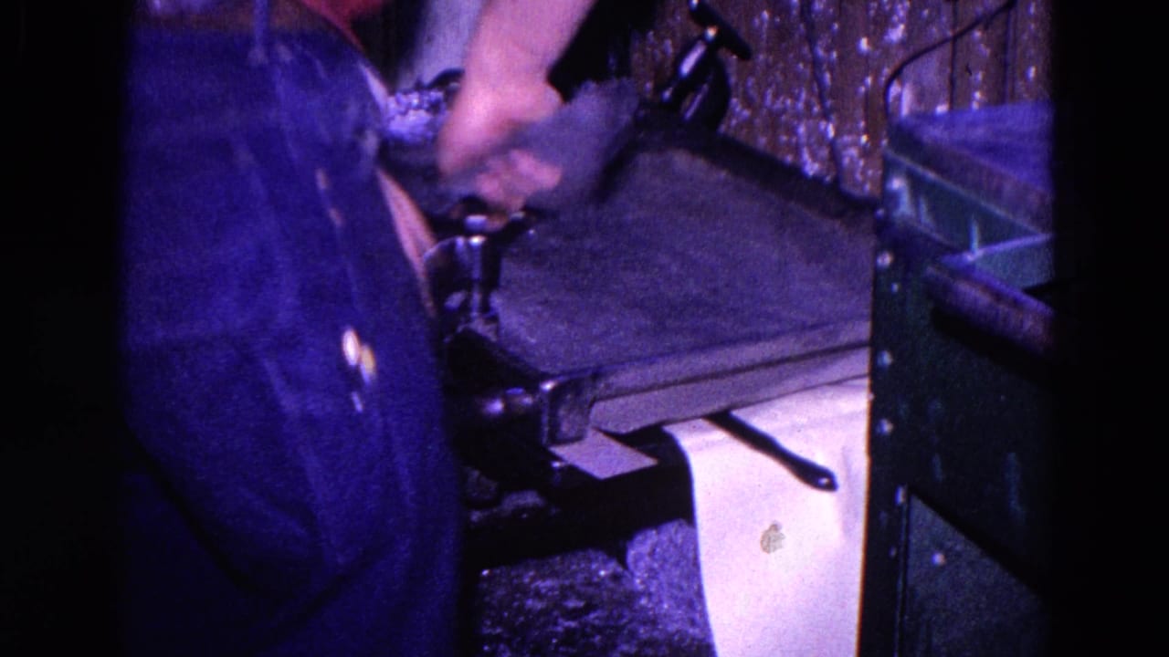 A person in dark clothes leans over the press, adjusting paper or the press mechanism in the Trenton workshop scene.
