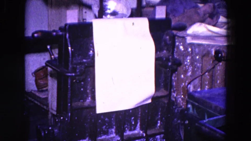 A sheet of paper is draped or fed through the metal press, with workshop tools and surfaces visible around it.