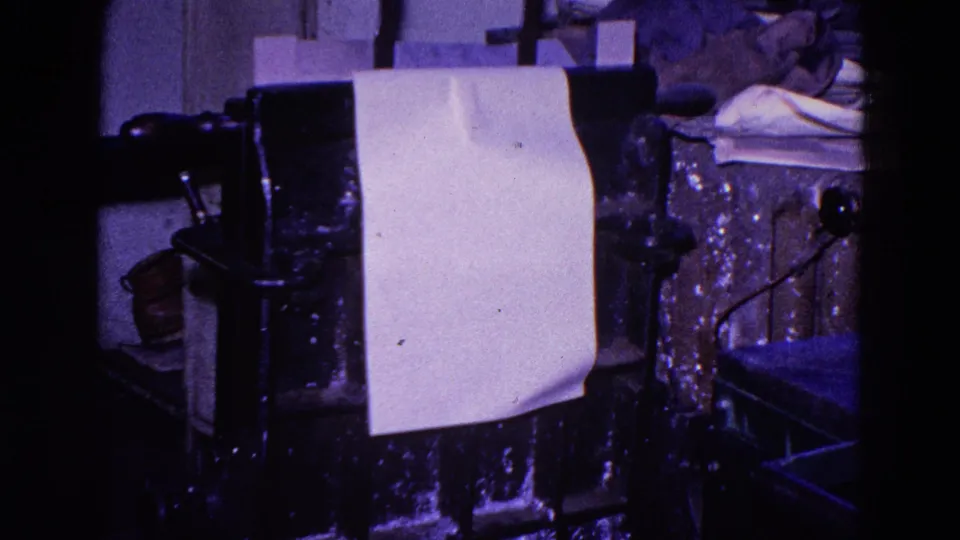The paper remains positioned on the metal press, seen with dark machinery, a handle, and a cluttered private workshop background.