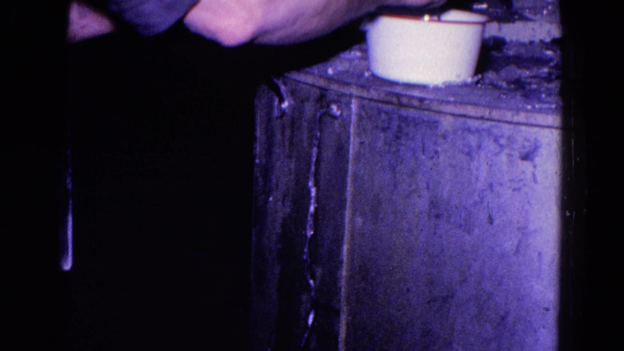 1967 Trenton home-movie frame showing hands near a white cup or small pot on a metal stove surface in dim interior light.