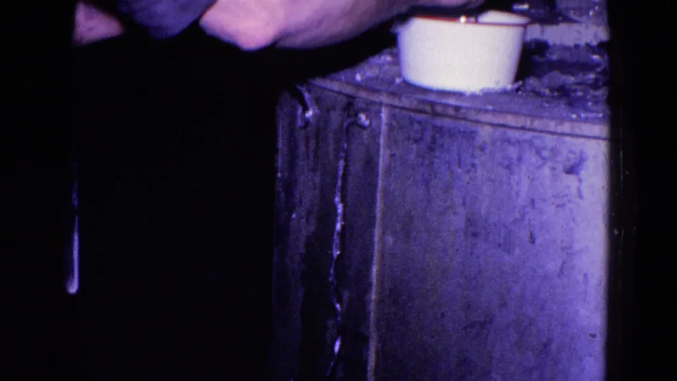 1967 Trenton home-movie frame showing hands near a white cup or small pot on a metal stove surface in dim interior light.