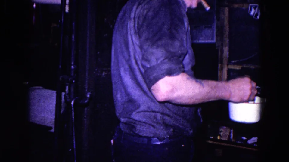 1967 Trenton home-movie frame showing an adult man in a dark shirt holding a white cup or small pot beside dark kitchen fixtures.
