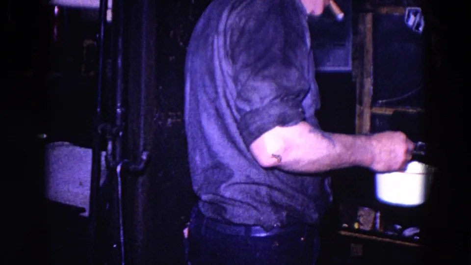 1967 Trenton home-movie frame with an adult man reaching toward a white cup or small pot in a shadowed private kitchen.