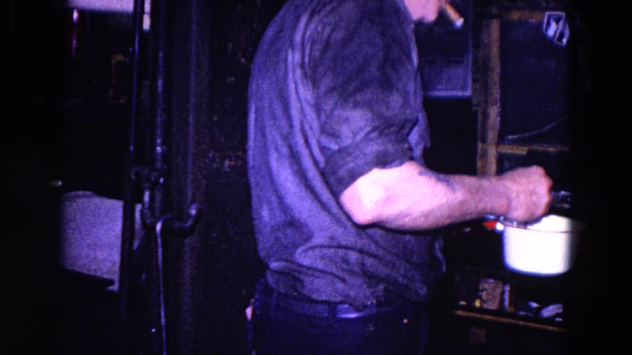 Man in a dark shirt stirring a white pot in a dim kitchen.