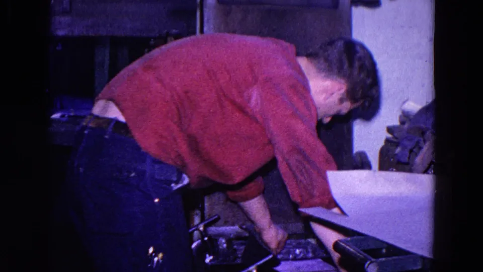 The 1967 Trenton home movie shows a person in a red sweater reaching near tools, papers, and a cluttered surface in a private interior.