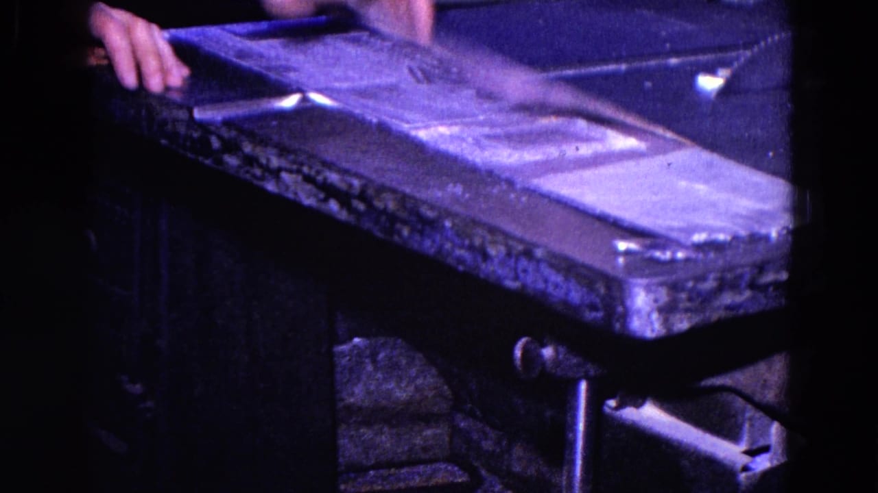 Worker's hands near a worn metal cutting table in a dark workshop.