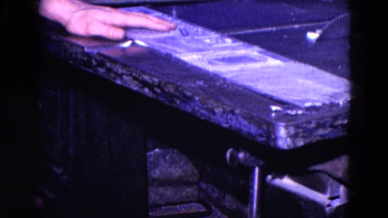 Close 8mm view from Trenton, New Jersey, with a worker's hand guiding material across a metal cutting table.