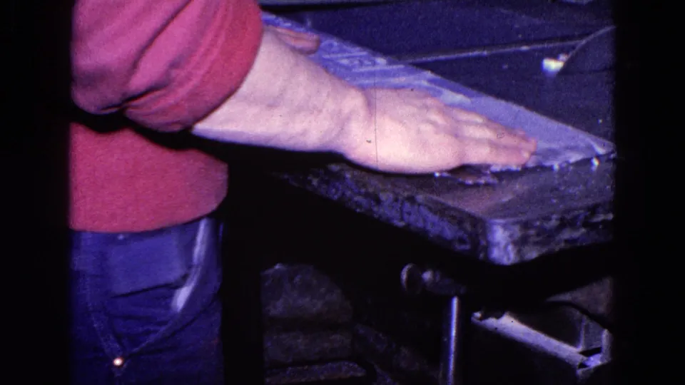 Worker in a red sweater and jeans leans over a cutting table in 1967 Trenton workshop footage.