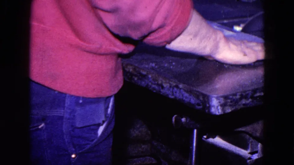 Close home-movie frame of a worker's red sweater, jeans, and outstretched arm at a workshop cutting machine in Trenton.