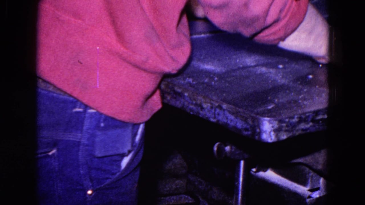 In 1967 Trenton footage, a person in a red sweater and blue jeans bends over a heavy metal machine in a dim industrial setting.