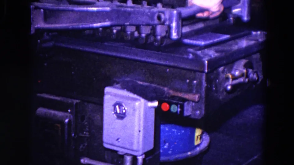 A hand reaches over metal tooling and controls on an industrial machine, with a gray electrical box and indicator lights below.