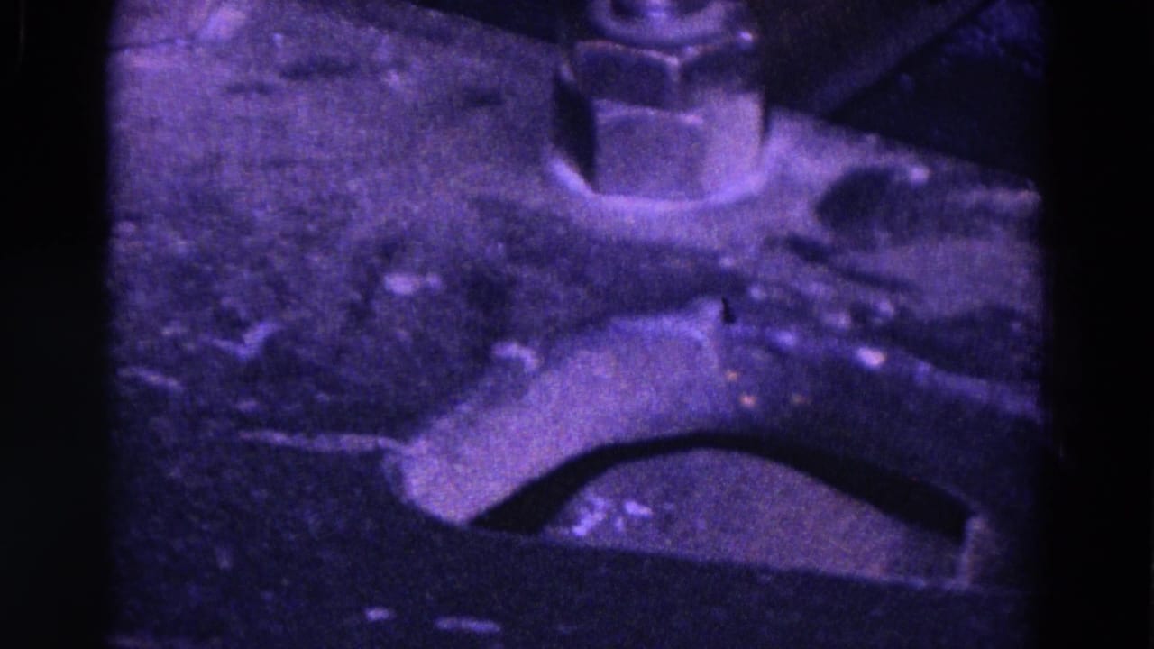Close-up from 1967 Trenton footage showing a curved industrial machine component, large bolt, and scuffed metal surface under purple-blue film color.