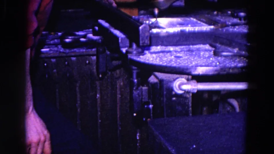 1967 home-movie frame showing a partial person’s arm near dark machinery-like surfaces and a metal bar under purple light.
